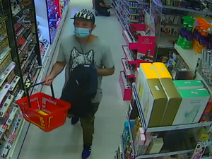 zdjęcie kolorowe: mężczyzna ubrany w szara koszulkę z krótki rękawie z wizerunkiem głowy wilka, trzymający granatowa bluzę dresową w ręce i czerwony koszyk sklepowy. Mężczyzna na głowie ma założoną czarno-biała czapkę z daszkiem a usta i nos zasłonięte niebieska maseczka ochronną