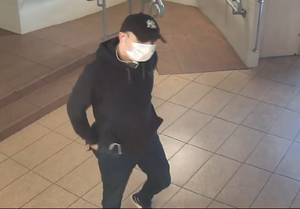 zdjęcie kolorowe: zrzut ekranu z zapisu monitoringu przedstawiającego szczupłego wysokiego mężczyznę ubranego w czarna czapkę z daszkiem z białym logo na przodzie, czarna bluzę dresową z kapturem, granatowe jeansy i czarne buty, który na twarzy ma założoną białą maseczkę ochronną