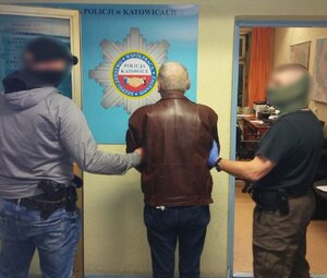 zdjęcie kolorowe: dwóch nieumundurowanych policjantów katowickiej komendy z zatrzymanym mężczyzną ubranym w brązową skórzaną kurtkę przed wejściem do pokoju