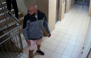 zdjęcie kolorowe: zapis z monitoringu na którym widoczny jest wysoki łysy mężczyzna ubrany w krótkie spodenki koloru różowego i szaro-czarna bluz z dresu z kapturem zapinaną na zamek