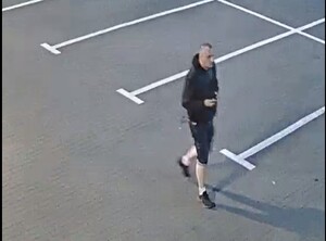 zdjęcie kolorowe: mężczyzna bardzo krótko obcięty ubrany w czarną rozpinaną bluzę dresową z kapturem, czarne krótkie spodenki, czarne buty sportowe i białe skarpety trzymający w prawej ręce telefon komórkowy