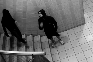 zdjęcie czarno-białe: mężczyzna ubrany w ciemne krótkie spodenki i ciemną bluzę dresową z kapturem założonym na głowę stojący na klatce schodowej