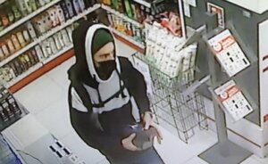 Zdjęcie kolorowe przedstawiające mężczyznę ubranego w biało-czarną bluzę z kapturem, z plecakiem i zieloną czapką na głowie stojącego w pomieszczeniu