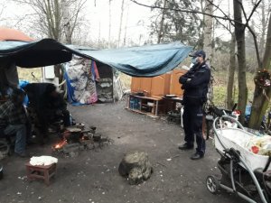 Zdjęcie kolorowe, na którym widać umundurowanego policjanta w miejscu przebywania bezdomnych na terenie leśnym. Na zdjęciu widać także osoby bezdomne.