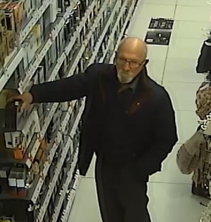 zdjęcie kolorowe: Mężczyzna w starszym wieku, łysina na czubku głowy, po bokach głowy siwe włosy, siwy zarost na brodzie, nosi okulary, na twarzy, ubrany w płaszcz koloru ciemnego – rozpięty, pod płaszczem widoczna górna część odzieży w kolorze ciemnym, spodnie z długimi nogawkami w kolorze ciemnym, buty koloru ciemnego, przez ramie posiadał przewieszoną torbę typu „aktówka” lub służącą do przenoszenia laptopa.