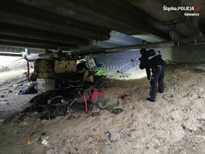 zdjęcie kolorowe: umundurowany policjant kontrolujący koczowisko osób bezdomnych umieszczone pod wiaduktem