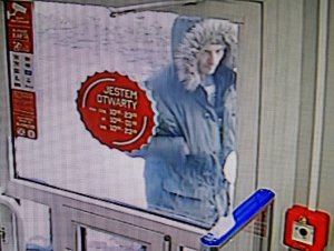 zdjęcie kolorowe: kadr z monitoringu sklepowego przedstawiający zniekształcony wizerunek mężczyzny stojącego za szklanymi drzwiami w kurtce z kapturem na głowie