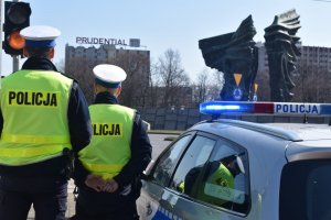 zdjęcie kolorowe: policjanci drogówki stojący przy radiowozie przed pomnikiem w centrum Katowic