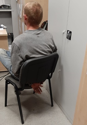 zdjęcie kolorowe: mężczyzna siedzący na krześle z rękami skutymi kajdankami trzymanymi z tyłu, ubrany w szara bluzę i niebieskie spodnie