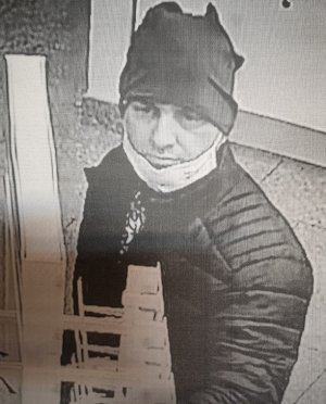 zdjęcie czarno-białe: mężczyzna podejrzewany o kradzież okularów, ubrany w ciemna kurtkę, ciemną czapkę i maseczkę ochronna założona na brodę