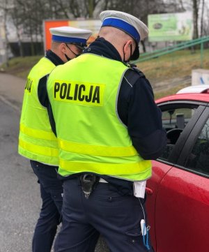 zdjęcie kolorowe: dwóch policjantów drogówki w zielonych kamizelkach odblaskowych z napisem Policja podczas kontroli czerwonego samochodu osobowego