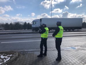 zdjęcie kolorowe: dwóch policjantów drogówki stojących przy krawędzi jezdni na tle samochodu ciężarowego typu TIR