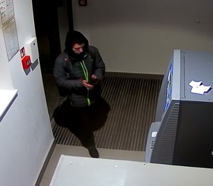 zdjęcie kolorowe: mężczyzna ubrany w ciemne spodnie, ciemną kurtkę z założonym kapturem na głowie wchodzący do pomieszczenia w którym ustawiony jest bankomat