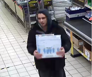 zdjęcie kolorowe: młody mężczyzna ubrany w ciemną bluzę dresową z założonym na głowę kapturem, ciemna kurtkę i ciemne spodnie, niosący biały karton