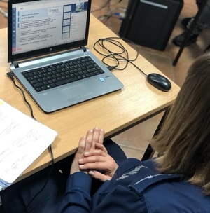 zdjęcie kolorowe: policjantka podczas prelekcji on-line, która siedzi na krześle przy stoliku  przed laptopem