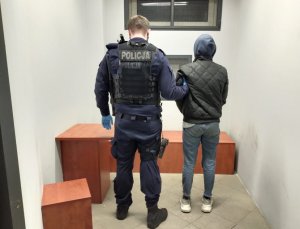 zdjęcie kolorowe: cela policyjnego aresztu w którym jest umundurowany policjant trzymający za ramię zatrzymana kobietę