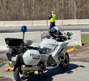 zdjęcie kolorowe: na pierwszym planie zaparkowany policyjny motocykl, a na drugim planie policjant katowickiej drogówki wykonujący pomiar prędkości
