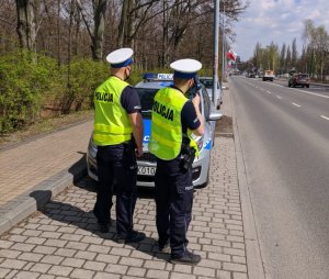 zdjęcie kolorowe: dwóch policjantów drogówki przy krawędzi jezdni, stojący przed oznakowanym radiowozem podczas działań &quot;Prędkość&quot;
