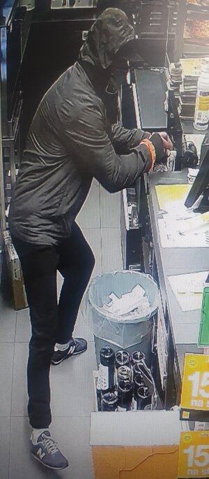 zdjęcie kolorowe: mężczyzna ubrany w czarne spodnie, ciemnoszarą kurtkę, czarną czapkę z daszkiem, niebieskie buty sportowe i czarna maseczkę założoną na twarzy, w chwili kradzieży gotówki z kasetki