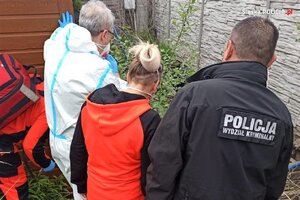 zdjęcie kolorowe: policjant ubrany w czarną kurtkę z napisem Policja Wydział Kryminalny, lekarz ubrany w biały ochronny kombinezon i maseczkę ochronna i ratowniczka medyczna w stroju służbowym