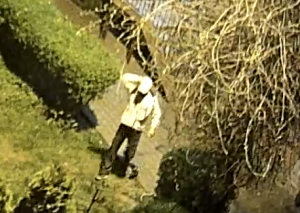 zdjęcie kolorowe: zrzut z monitoringu przedstawiający mężczyznę w ciemnych spodniach i jasnej bluzie z założoną białą czapką stojącego na skwerku przed blokiem
