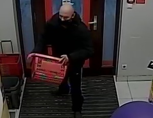 zdjęcie kolorowe: zrzut ze sklepowego monitoringu przedstawiający łysego mężczyznę ubranego w czarne buty, czarne spodnie, czarna kurtkę i czarna maseczkę ochronna na twarzy niosący czerwony sklepowy koszyk zakupowy