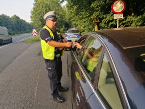 zdjęcie kolorowe: policjant wydziału ruchu drogowego przekazujący zawieszkę kierującemu czarnym samochodem osobowym