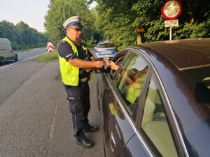 zdjęcie kolorowe: policjant wydziału ruchu drogowego przekazujący zawieszkę kierującemu czarnym samochodem osobowym
