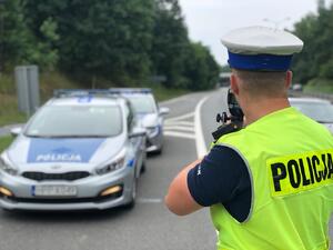 zdjęcie kolorowe: policjant katowickiej drogówki na tle dwóch radiowozów wykonujący pomiar prędkości