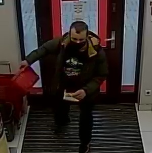 zdjęcie kolorowe: zrzut z monitoringu sklepowego przedstawiający mężczyznę w ciemnozielonej kurtce, ciemnych spodniach i czarnych butach z maseczka ochronna na twarzy wchodzącego do sklepu