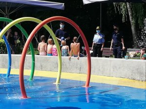 Zdjęcie kolorowe, na którym widać dzieci siedzące tyłem, funkcjonariuszy policji, strażnika miejskiego, a także infrastrukturę wodnego placu zabaw.