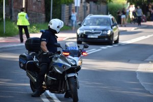 Zdjęcie kolorowe przedstawia policjanta na motocyklu, na tle pojazdu na trasie 78. Tour de Pologne.
