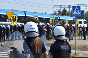 Zdjęcie kolorowe, na którym widać radiowóz policyjny oraz policjantów zabezpieczających przejście kibiców.