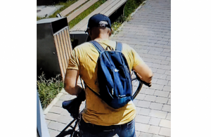 zdjęcie kolorowe: mężczyzna stojący tyłem do kamery ubrany w żółtą koszulkę z krótkim rękawem, niebieskie spodnie i niebieski plecak założony na ramiona oraz czarna czapkę z daszkiem, trzymający rower