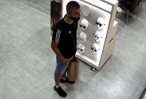 zdjęcie kolorowe: mężczyzna znajdujący się w salonie optycznym, ubrany w krótkie jeansowe spodnie i czarna koszulkę z trzema białymi paskami na ramieniu, z papierowa torba w ręce i czarna maseczka ochronna założoną pod brodą