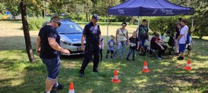 zdjęcie kolorowe: dzielnicowy przyglądający się męzczyźnie w alkogoglach, który pokonuje tor przeszkód w towarzystwie innych uczestników festynu