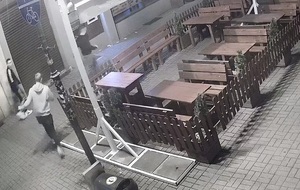 zdjęcie czarno-białe: zrzut ekranu z nagrania monitoringu przedstawiające 3 młodych mężczyzn w rejon ogródka restauracyjnego