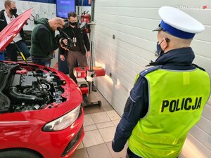 zdjęcie kolorowe: policjant wydziału ruchu drogowego na stacji diagnostycznej przyglądający się pracy diagnosty, w tle dwóch operatorów kamer telewizyjnych