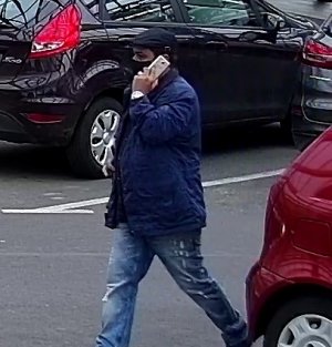 zdjęcie kolorowe: mężczyzna w czarnym kaszkiecie, granatowej kurtce z czarna maseczka ochronna na twarzy rozmawiający na parkingu przez telefon komórkowy