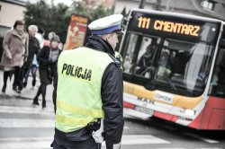 zdjęcie kolorowe: policjant drogówki w rejonie przejścia dla pieszych i przystanku komunikacji miejskiej, w tle autobus komunikacji miejskiej