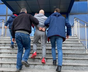 zdjęcie kolorowe: na schodach przed wejściem do Komendy Miejskiej Policji w Katowicach policjanci wydziału kryminalnego wprowadzają zatrzymanego mężczyznę poszukiwanego europejskim nakazem aresztowania, któremu założono kajdanki zespolone