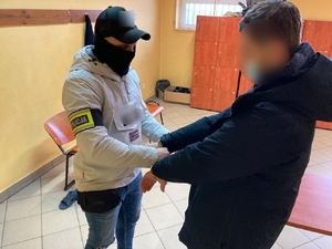 zdjęcie kolorowe: policjant wydziału kryminalnego zakłada kajdanki zatrzymanemu mężczyźnie na ręce trzymane z przodu