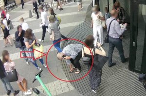 zdjęcie kolorowe: mężczyzna w średnim wieku, wysoki, w krótkich siwych włosach ubrany w jasną koszulę w kratę z długim rękawem, czarne spodnie do kostek z materiału i ciemne klapki &#039;&#039;japonki&#039;&#039; schyla się i podnosi kamerę z ziemi przed galerią handlową