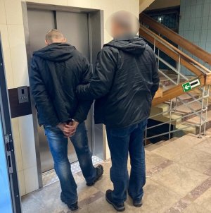 zdjęcie kolorowe: korytarz komendy przed windą, zatrzymany mężczyzna z kajdankami założonymi na ręce trzymane z tyłu trzymany za ramię przez policjanta wydziału kryminalnego katowickiej komendy