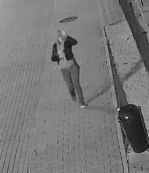 zdjęcie czarno białe: zrzut z monitoringu przedstawiający osobę idącą chodnikiem, ubraną w ciemne spodnie, ciemną bluzę z kapturem założoną na głowę i czarna kurtkę,
