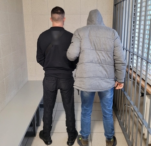 zdjęcie kolorowe: zatrzymany mężczyzna podejrzany o kierowanie gróźb karalnych wobec trzech osób, w pomieszczeniu dla osób zatrzymanych razem z trzymającym go za ramię policjantem