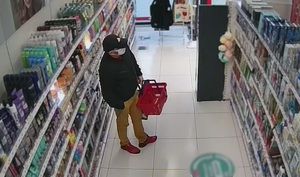 zdjęcie kolorowe: wysoki mężczyzna ubrany w musztardowe spodnie, ciemną kurtkę, czarna czapkę z daszkiem z założoną maseczka ochronna na twarzy trzymający czerwony koszyk sklepowy