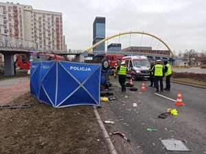 zdjęcie kolorowe: policyjny parawan z napisem Policja zasłaniający samochód, 4 policjantów ruchu drogowego obsługujących zdarzenie, karetka pogotowia i wóz strażacki