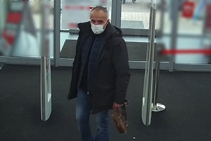 zdjęcie kolorowe: mężczyzna w średnim wieku ubrany w granatowe jeansy, czarna kurtkę z maseczką ochronna na twarzy wchodzący do marketu