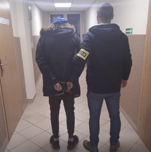 zdjęcie kolorowe:  mężczyzna ubrany w ciemne spodnie i ciemną długa kurtkę z kapturem, który jest trzymany za rękę przez policjanta kryminalnego